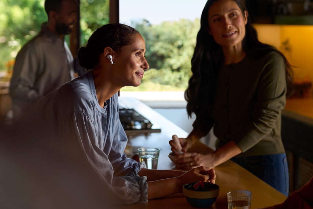 Person in the center showing new AirPods, there is a person in the back, as well as another person to the right, showing a user in context