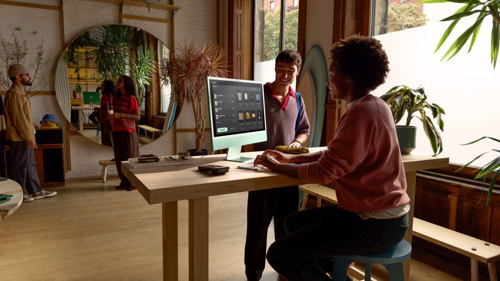 Two people using a green iMac on a desk in a studio with two more people in the background talking to each other